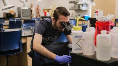 A student wearing blue latex gloves looks into a microscope.