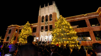 The pine trees in front of Weller Hall lit up with Christmas lights as students and community sing Christmas carols