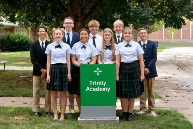 9 high school students in uniform stand around a sign that reads Trinity Academy.