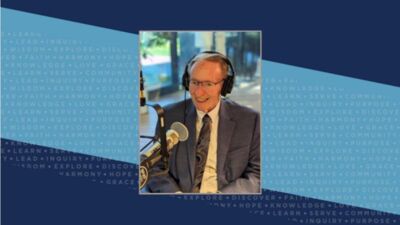 An older gentleman in a grey suit and swirly tie sits in front of a radio station microphone. He is also wearing a headset and glasses.