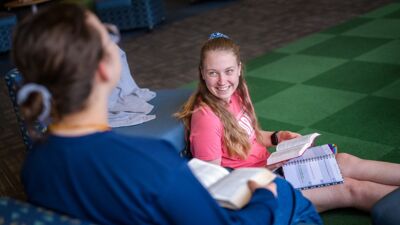 A student sitting on the carpet smiling, reading the Bible with her friend