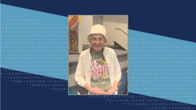 An older woman smiles at the camera. She has white hair and is wearing a white bucket hat and a t-shirt that says 