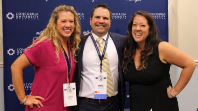 Three Concordia Nebraska alumni posing in front of a backdrop with the university logo