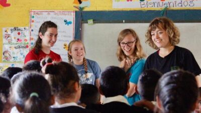 Four college aged women stand at the front of a classroom. They are smiling and laughing. The backs of their students' heads can be seen. Above the teachers is a hand-lettered sign reading 